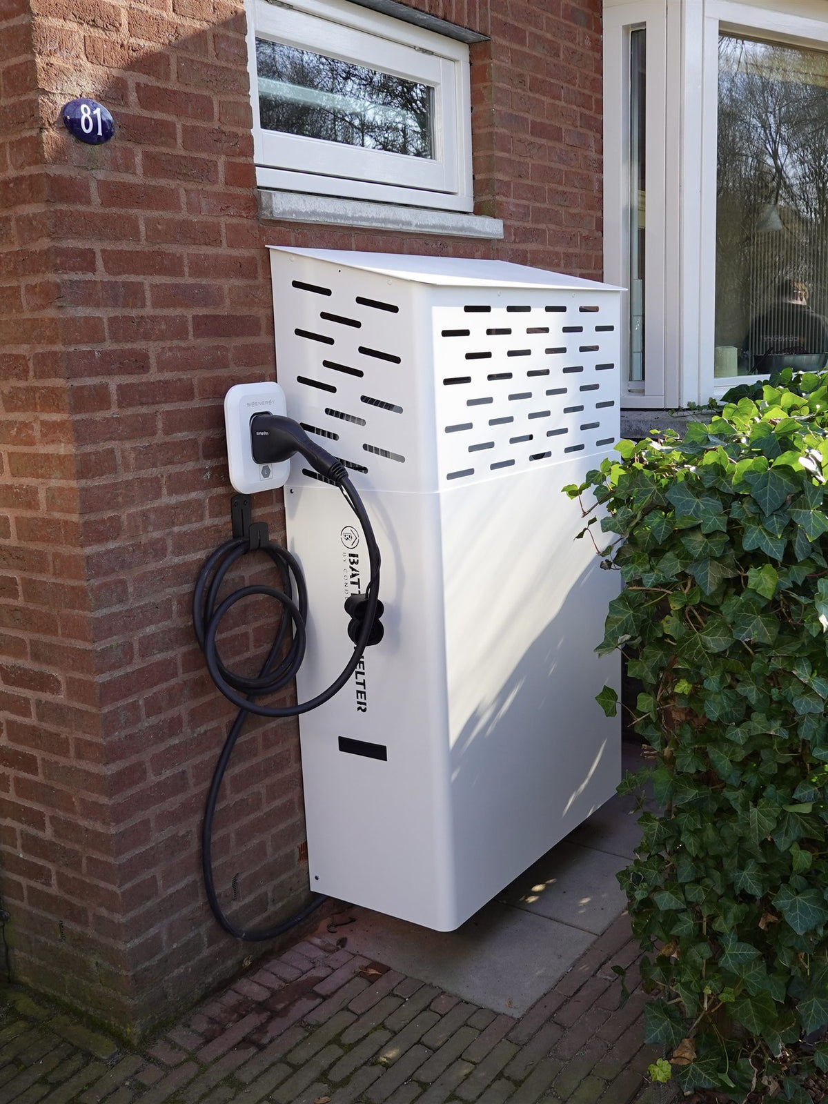 Witte Batteryshelter aan een bakstenen gevel naast de voordeur, met laadpaal